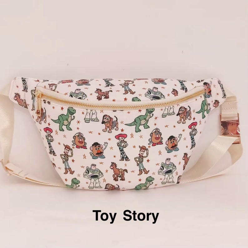 May include: White fanny pack with a gold zipper and strap. The bag is covered in colorful cartoon characters from the movie "Toy Story". The strap is a cream color. The text "Toy Story" is at the bottom of the image.