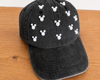 May include: Black denim baseball cap adorned with white pearl-like Mickey Mouse head shapes. The cap has a curved brim and a button on top. The hat is sitting on a wooden surface.