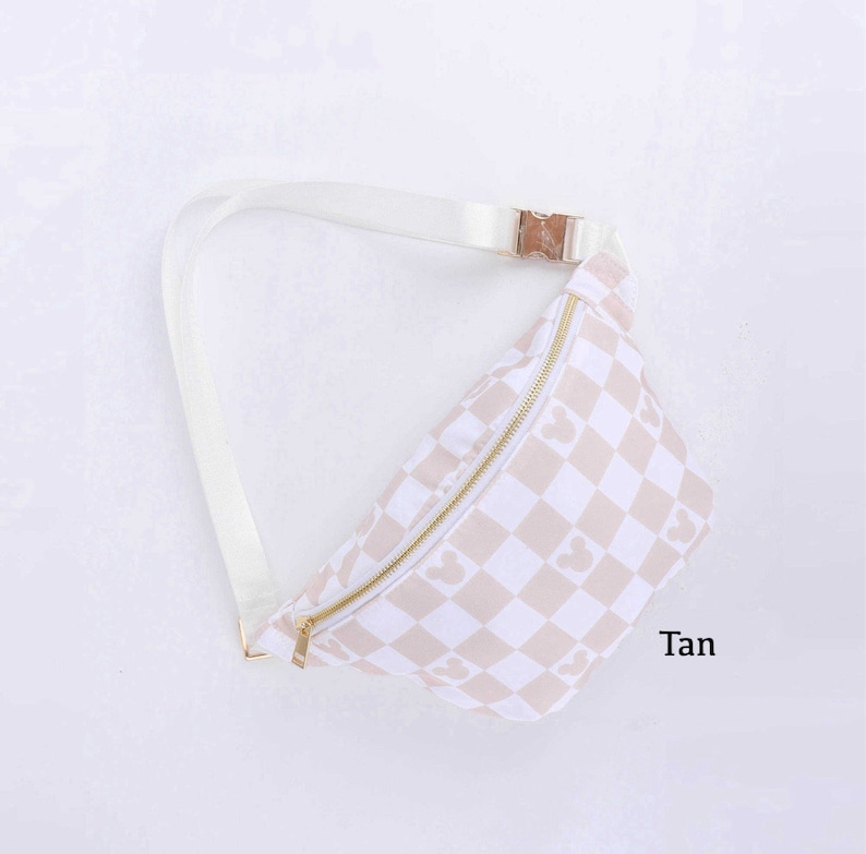 May include: Tan fanny pack with a checkered pattern featuring Mickey Mouse silhouettes. The bag has a gold zipper and buckle, and a white adjustable strap. The word "Tan" is printed on the lower right corner.