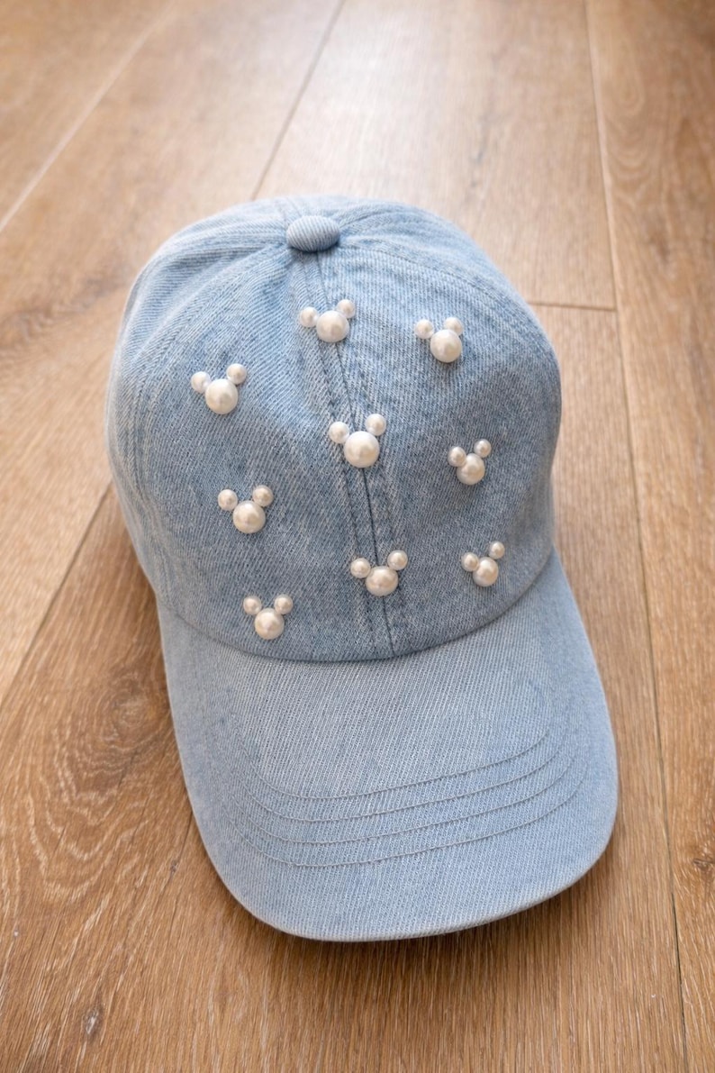 May include: Light blue denim baseball cap adorned with white pearl-like embellishments arranged in a repeating pattern. The cap features a curved brim and a button on top. A stylish accessory for casual wear.