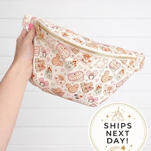 May include: A cream-colored fanny pack featuring a repeating pattern of brown and pink line-art illustrations including cameras, tickets, mouse ears, and castle icons. The bag has a gold-tone zipper closure and an adjustable strap for waist wear.