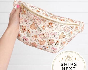 May include: A cream-colored fanny pack featuring a repeating pattern of brown and pink line-art illustrations including cameras, tickets, mouse ears, and castle icons. The bag has a gold-tone zipper closure and an adjustable strap for waist wear.
