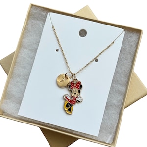 May include: Gold-toned necklace featuring a Minnie Mouse pendant with red, black, and yellow enamel detailing. The necklace includes a small, round charm with a letter. Presented in a gold gift box with a white display card.