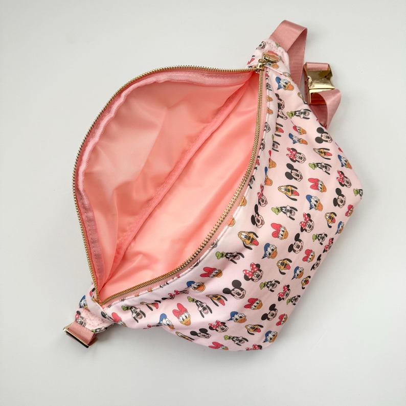 May include: Pink fanny pack with a gold zipper and buckle. The bag is patterned with cartoon character faces. The interior is a solid pink color. The strap is pink.