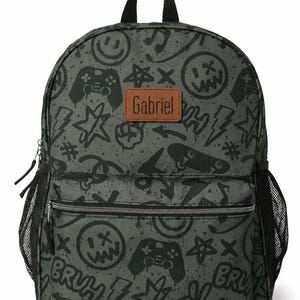 Op de afbeelding: Een donkergrijze rugzak met een zwarte graffiti-print van smileys, gamecontrollers, bliksemschichten, sterren en het woord BRUH. Een bruine rechthoekige patch met de naam Gabriel is in het midden op de voorkant boven een ritsvak geplaatst.
