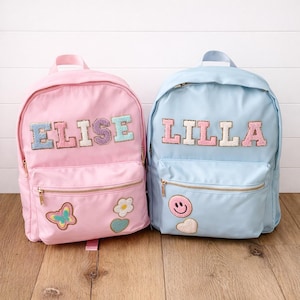 May include: Two personalized backpacks, one pink and one light blue, with gold zippers. The pink backpack has the name "ELISE" in colorful, textured letters, and patches of a butterfly, flower, and heart. The blue backpack says "LILLA" and has a smiley face and heart patch.