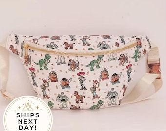 May include: A white fanny pack featuring an all-over print of various cartoon characters including a cowboy, a space ranger, a green dinosaur, and a potato-shaped figure, accented with small gold stars and a gold-toned metal zipper closure.