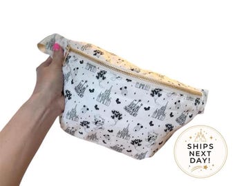 May include: A white fanny pack featuring a black line-art pattern of castles, balloons, and character silhouettes. The bag has a gold-toned zipper closure along the top edge. The fabric is printed with repeating icons and the text It's a small world.