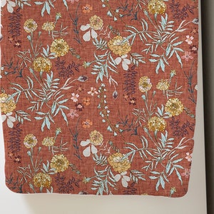 May include: A rectangular piece of fabric with a floral pattern. The fabric has a reddish-brown background with yellow, white, and pink flowers, and light blue leaves. The design is detailed and appears to be printed on the fabric.