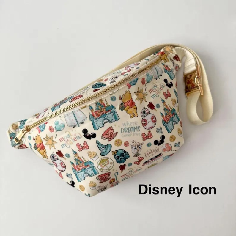 May include: Cream-colored fanny pack with a gold zipper and strap. The bag is patterned with Disney character icons, including castles, Winnie the Pooh, and Mickey Mouse. The text "Disney Icon" is printed on the bottom right.