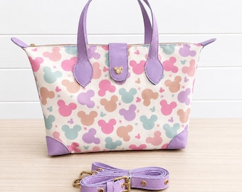 May include: A white tote bag with a colorful Mickey Mouse pattern, featuring pink, blue, and peach-colored silhouettes. The bag has purple handles, trim, and a detachable shoulder strap. A gold-colored clasp secures the top flap.