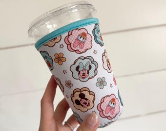 May include: A white neoprene cup sleeve featuring a repeating pattern of cartoon characters including Mickey Mouse, Minnie Mouse, and Daisy Duck inside scalloped floral shapes, accented with small stars and flowers, finished with a bright blue trim.