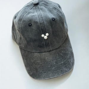 May include: A gray baseball cap with a distressed finish. The cap features a small button on top and three white pearl embellishments below the brim. Two small black embellishments are on either side of the cap.