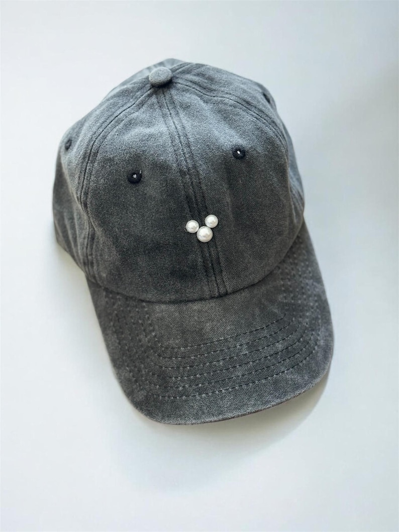 May include: A gray baseball cap with a distressed finish. The cap features a small button on top and three white pearl embellishments below the brim. Two small black embellishments are on either side of the cap.