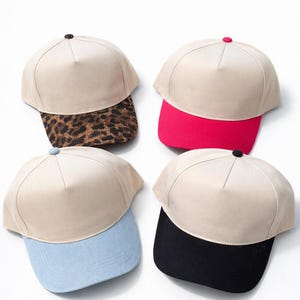 May include: Four trucker hats with beige crowns and different colored brims. One has a leopard print brim, another a bright pink brim, a light blue brim, and a black brim. The text "Trucker Hat Options" is at the top.