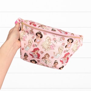 May include: Pink fanny pack featuring a repeating pattern of cartoon princesses and roses. The bag has a gold zipper and a pink zipper pull. The fanny pack is held by a hand.
