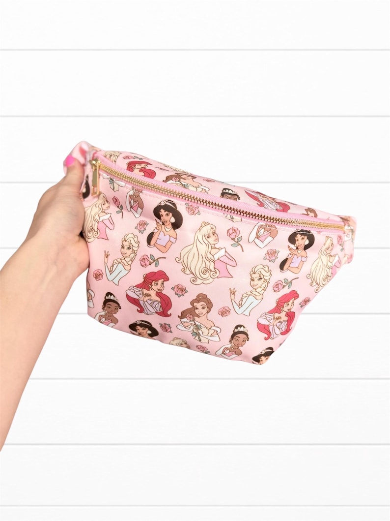 May include: Pink fanny pack featuring a repeating pattern of cartoon princesses and roses. The bag has a gold zipper and a pink zipper pull. The fanny pack is held by a hand.