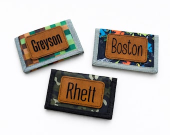 May include: Three rectangular wallets with grey trim, each featuring a brown leather-like patch centered on the front. The wallets display different patterns: green plaid, colorful dinosaur print, and dark camouflage, with the names Greyson, Boston, and Rhett.