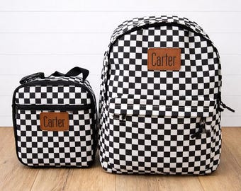 May include: A black and white checkered backpack and matching lunch bag. Both items have a brown leather patch with the name "Carter" on them. The backpack has a front pocket and adjustable straps. The lunch bag has a handle and a zipper closure.