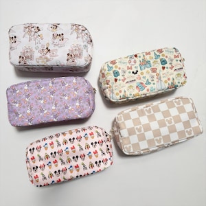 May include: Five rectangular cosmetic bags with zipper closures. The bags feature various patterns, including Mickey Mouse, Daisy Duck, and a checkered design with Mickey Mouse silhouettes. Colors include lavender, pink, and cream.
