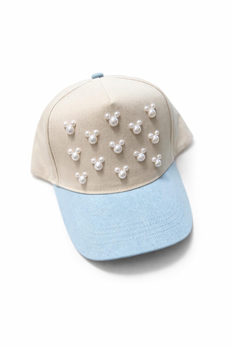 May include: A beige baseball cap with a light blue brim. The cap is decorated with multiple pearl-like embellishments arranged in a Mickey Mouse head shape.