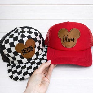 May include: Two baseball caps with Mickey Mouse ear-shaped patches. One cap is red with a mesh back and the name "Olivia" in script. The other is black and white checkered with the name "Greyson".