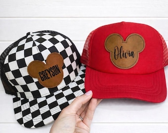 May include: Two baseball caps with Mickey Mouse ear-shaped patches. One cap is red with a mesh back and the name "Olivia" in script. The other is black and white checkered with the name "Greyson".