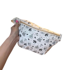May include: White fanny pack with a gold zipper and a black and white pattern featuring castles, balloons, and the text "small world". The bag is held by a hand and has a curved shape.