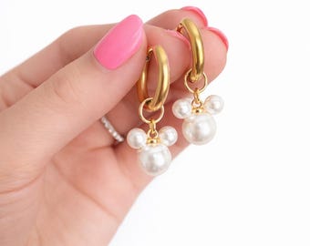 May include: A pair of gold hoop earrings with a cluster of white faux pearls. The earrings are held up by a hand with pink nail polish. The earrings have a classic and elegant design.