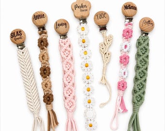 Puede incluir: Una colección de broches para chupetes de bebé hechos a mano en varios diseños de macramé. Cada broche tiene una parte superior de madera con un nombre grabado. Los broches son en tonos blanco, marrón, rosa y verde, con detalles de margaritas y flores.