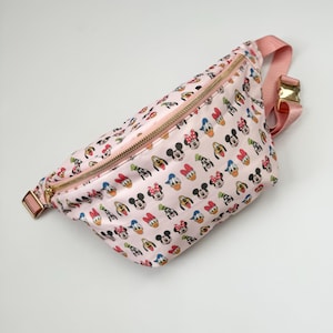 May include: A light pink fanny pack featuring an all-over print of various Disney character faces including Mickey Mouse, Minnie Mouse, Donald Duck, and Goofy. The bag has a gold-toned metal zipper closure and an adjustable pink strap with a gold buckle.