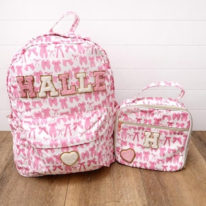 May include: A pink and white backpack and matching lunch bag set. Both items feature a repeating pattern of pink bows and ballet shoes. The backpack is personalized with the name "HALLE" in pink and gold letters, and the lunch bag has the letter "H".