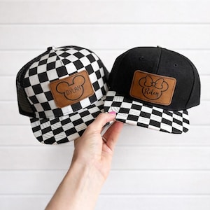 May include: Two baseball caps, one black and one with a black and white checkerboard pattern. Both have a brown leather patch with a cartoon mouse head design. The black cap says "Riley" and the checkerboard cap says "Dylan".
