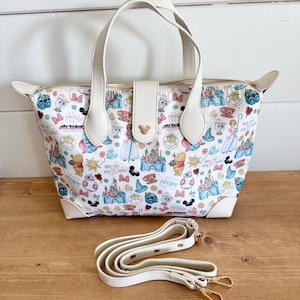May include: White tote bag with a colorful pattern featuring cartoon castles, characters, and text. The bag has a white handle, a detachable shoulder strap, and gold-colored hardware. The bag is sitting on a wooden surface.