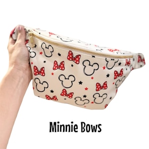 May include: Cream-colored fanny pack with a gold zipper, featuring a pattern of black Mickey Mouse heads, red polka-dot bows, and black stars. The text "Minnie Bows" is printed in black at the bottom.