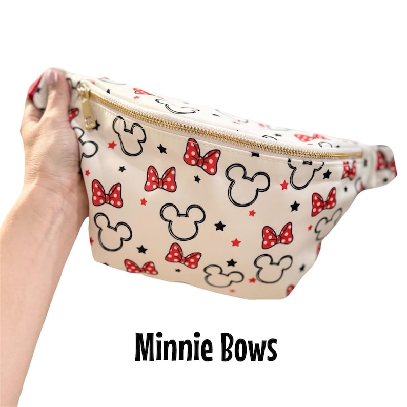 May include: Cream-colored fanny pack with a gold zipper, featuring a pattern of black Mickey Mouse heads, red polka-dot bows, and black stars. The text "Minnie Bows" is printed in black at the bottom.