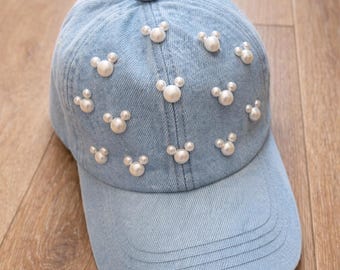 May include: A light blue denim baseball cap featuring multiple white pearl embellishments arranged in the shape of small mouse heads. The cap has a structured crown, a curved brim with stitched detailing, and a matching fabric button on top.