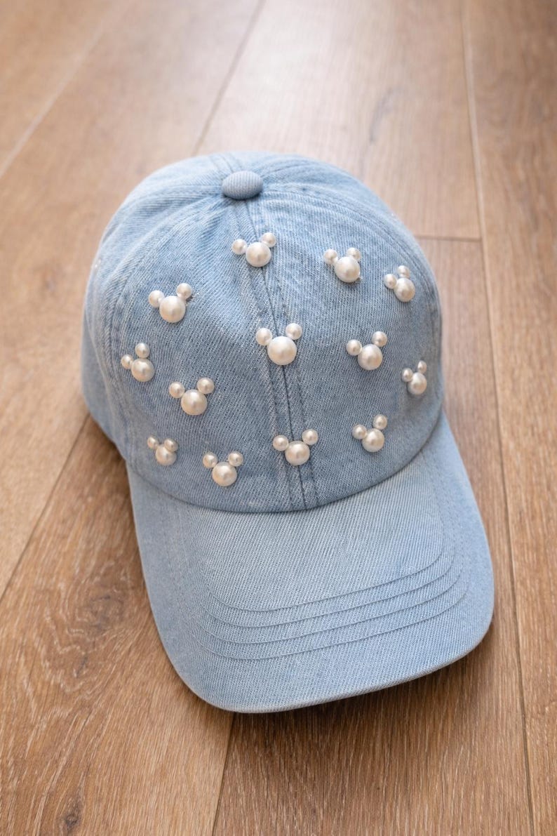 May include: Light blue denim baseball cap adorned with white pearl-like Mickey Mouse head shapes. The cap features a curved brim and a button on top. The hat is set against a wooden background.