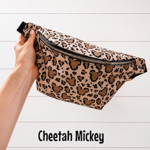 May include: A tan and brown cheetah print fanny pack with black trim and a black zipper. The fanny pack features brown Mickey Mouse head shapes within the cheetah print. The text "Cheetah Mickey" is displayed below the fanny pack.