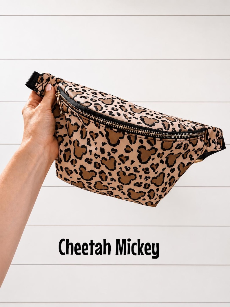 May include: A tan and brown cheetah print fanny pack with black trim and a black zipper. The fanny pack features brown Mickey Mouse head shapes within the cheetah print. The text "Cheetah Mickey" is displayed below the fanny pack.