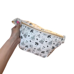 May include: White fanny pack with a gold zipper and a black and white pattern featuring castles, balloons, and the words "Small World". The bag is held by a person.