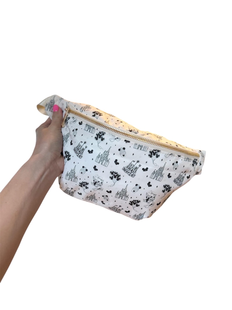May include: White fanny pack with a gold zipper and a black and white pattern featuring castles, balloons, and the words "Small World". The bag is held by a person.
