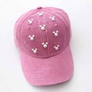 May include: A pink baseball cap adorned with white pearl embellishments arranged in a Mickey Mouse-inspired pattern. The cap features a curved brim and a fabric-covered button on top.