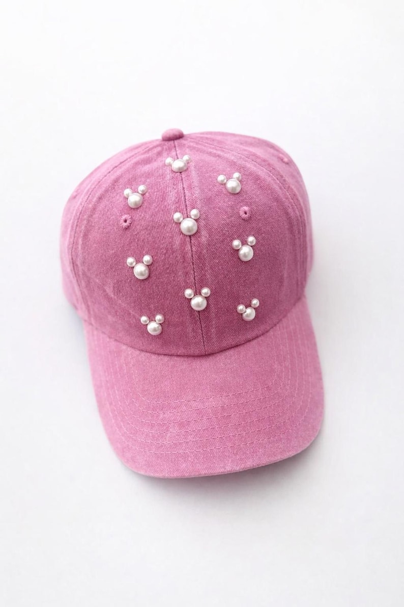 May include: A pink baseball cap adorned with white pearl embellishments arranged in a Mickey Mouse-inspired pattern. The cap features a curved brim and a fabric-covered button on top.