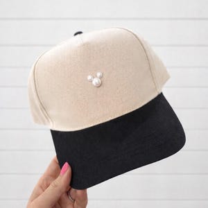 May include: A beige and black baseball cap with a pearl embellishment. The cap features a beige crown and a black brim. Three pearls are arranged on the front of the cap. The cap is held by a person.