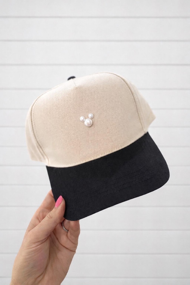 May include: A beige and black baseball cap with a pearl embellishment. The cap features a beige crown and a black brim. Three pearls are arranged on the front of the cap. The cap is held by a person.