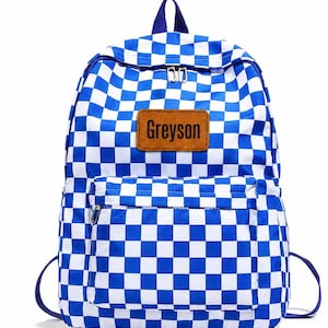 May include: A blue and white checkered backpack featuring a rectangular brown leather-like patch on the front pocket with the name Greyson printed in black text. The bag includes a top handle, two shoulder straps, and silver-toned metal zippers.