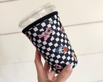 May include: A black and white checkered neoprene cup sleeve featuring a repeating pattern of cartoon characters including Minnie Mouse, Donald Duck, and Pluto, topped with a clear plastic lid on a beverage container.