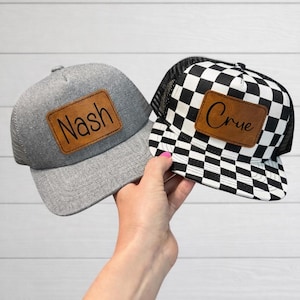 May include: Two trucker hats. One is grey with a mesh back and a brown leather patch that says "Nash". The other is black and white chequered with a mesh back and a brown leather patch that says "Crue".