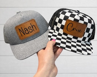 May include: Two trucker hats. One is gray with a mesh back and a brown leather patch that says "Nash". The other is black and white checkered with a mesh back and a brown leather patch that says "Crue".